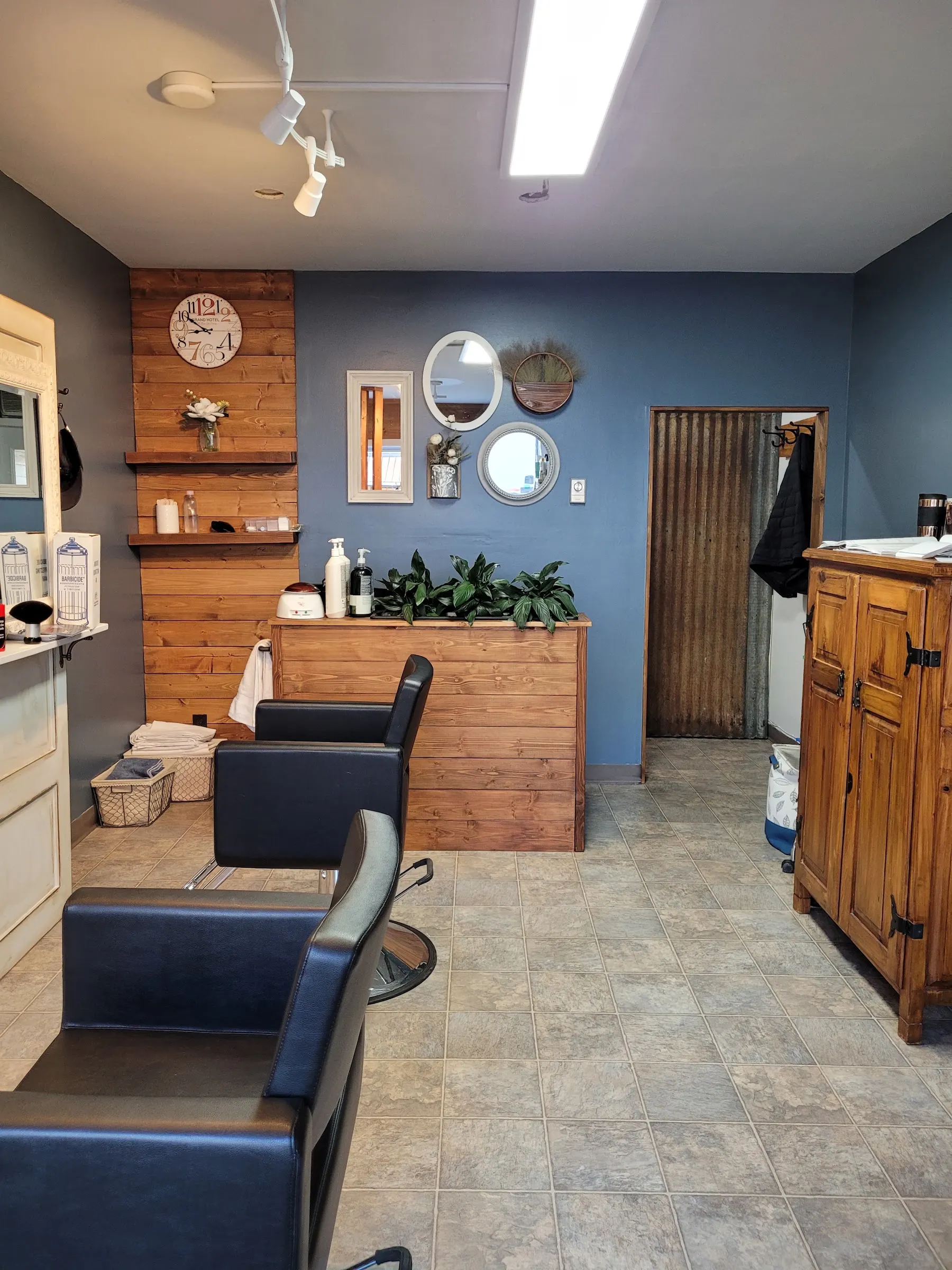 Inside Mane Obsession Salon: slate-blue walls, a stained pine plank wall with a station clock, black styling chairs and peace lilies on the counter