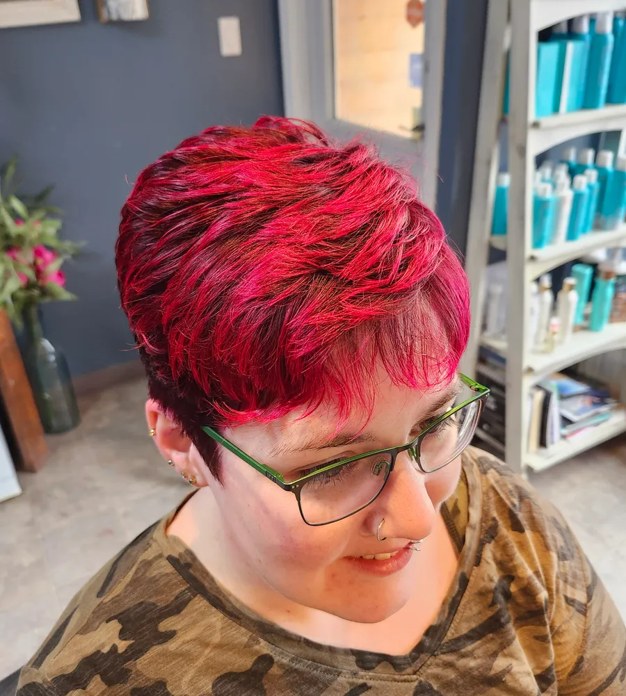 A textured pixie cut in vivid magenta-red, worn with green-framed glasses